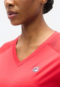 Κόκκινο αθλητικό πουκάμισο με V λαιμόκοψη, κατασκευασμένο από απαλό ύφασμα. Διαθέτει ένα μικρό μπλε και λευκό λογότυπο στην αριστερή πλευρά και υφασμάτινες λεπτομέρειες στους ώμους.