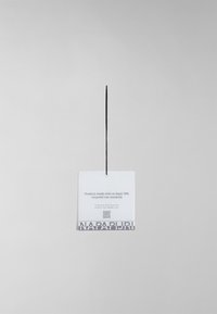 Etichetta bianca sospesa con testo nero, caratterizzata da una nota sui materiali riciclati e un codice QR, accentuata da un logo "NAPAPIJRI" grigio in basso.