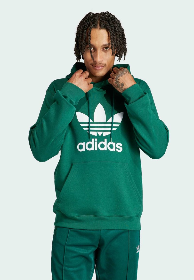 Zelená mikina s kapucňou vyrobená z mäkkej tkaniny, s veľkým bielym logom Adidas a klokania kapsou. Skombinujte s zelenými nohavicami.