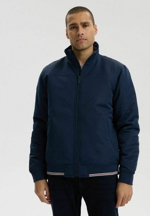 Mężczyzna ubrany w granatową kurtkę na zamek z pionowo stojącym kołnierzem i paskami przy dolnym brzegu, stojący na jednolitym tle.