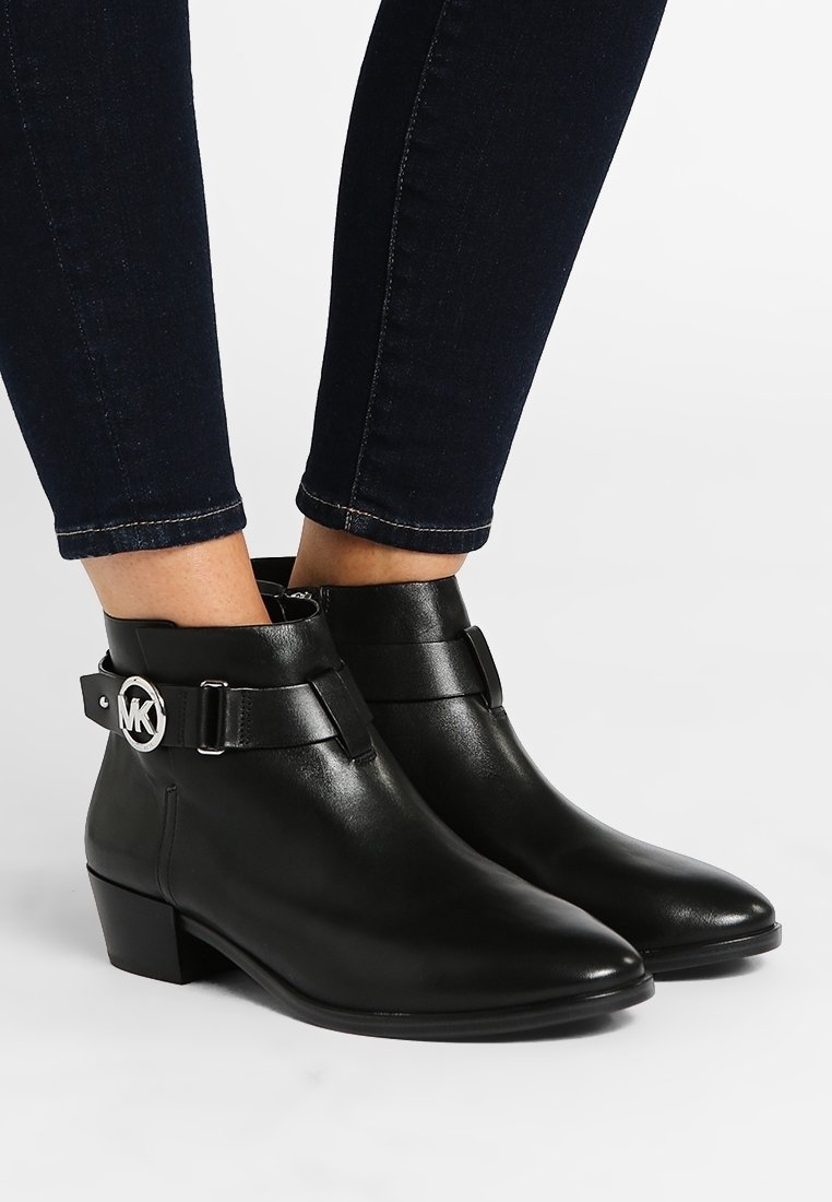 Bottines en cuir noir avec talons bas, bouts pointus et sangles latérales ornées de logos circulaires argentés, portées avec un jean skinny foncé.