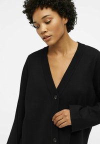 Zwarte oversized cardigan van ribgebreid stof, met een diepe V-hals, knopen aan de voorkant en wijde mouwen. Soepele textuur en minimalistisch ontwerp.
