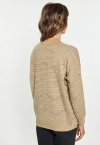 Beige kötött pulóver laza szabásban, vízszintes csíkokkal, lyukacsos részletekkel és bordázott aljjal. Puha textúra és lezser sziluett.