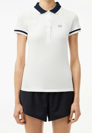 Kvinde iført en hvid poloshirt med korte ærmer og navy krave og kant, kombineret med sorte shorts, stående med armene afslappet ned langs siderne.