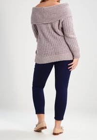 Schulterfreier, gerippter Pullover in hellem Mauve, mit lockerem Schnitt und strukturiertem Strick, kombiniert mit dunklen marineblauen, eng geschnittenen Hosen und hellen Schuhen.