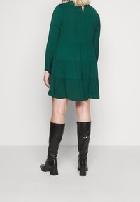 Vestito verde a strati con maniche lunghe e dettaglio a chiave sulla schiena, abbinato a stivali neri in pelle al ginocchio. Tessuto liscio, design casual.