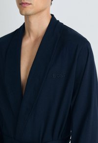 Temno modra bombažna kopalna plašča z ovratnikom v stilu šal, opremljena z izvezenim logotipom "BOSS" na levem prsnem delu in pasom s pasom. Mehka tekstura, srednje dolžine.
