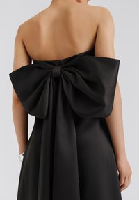 Vestido de satén negro con un gran lazo en la espalda, textura suave, corsé ajustado y falda fluida con un sutil brillo.