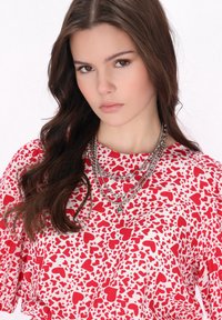 Bluză roșie și albă cu un model în forme mici de inimă, cu mâneci scurte și coliere din lanțuri de argint suprapuse. Textură netedă, design casual.