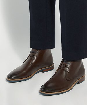 Bottines en cuir marron avec lacets noirs portées avec un pantalon foncé sur un fond de sol clair.