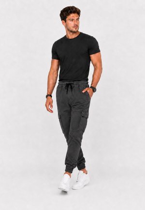Mann geht, trägt schwarzes T-Shirt, dunkelgraue Cargo-Jogginghosen, weiße Sneakers und eine schwarze Uhr am linken Handgelenk.