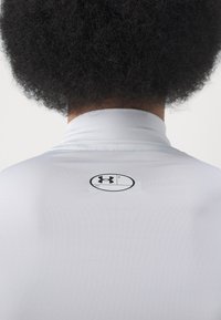 Biała koszulka sportowa z wysokim kołnierzem, z czarnym logo Under Armour z tyłu i gładką, elastyczną fakturą materiału.