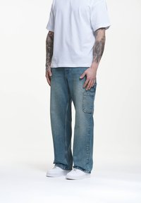 Jeans de mezclilla azul claro con un corte holgado, que cuentan con un bolsillo lateral y un ligero desgaste. Combinados con una camiseta blanca de manga corta y zapatillas blancas.