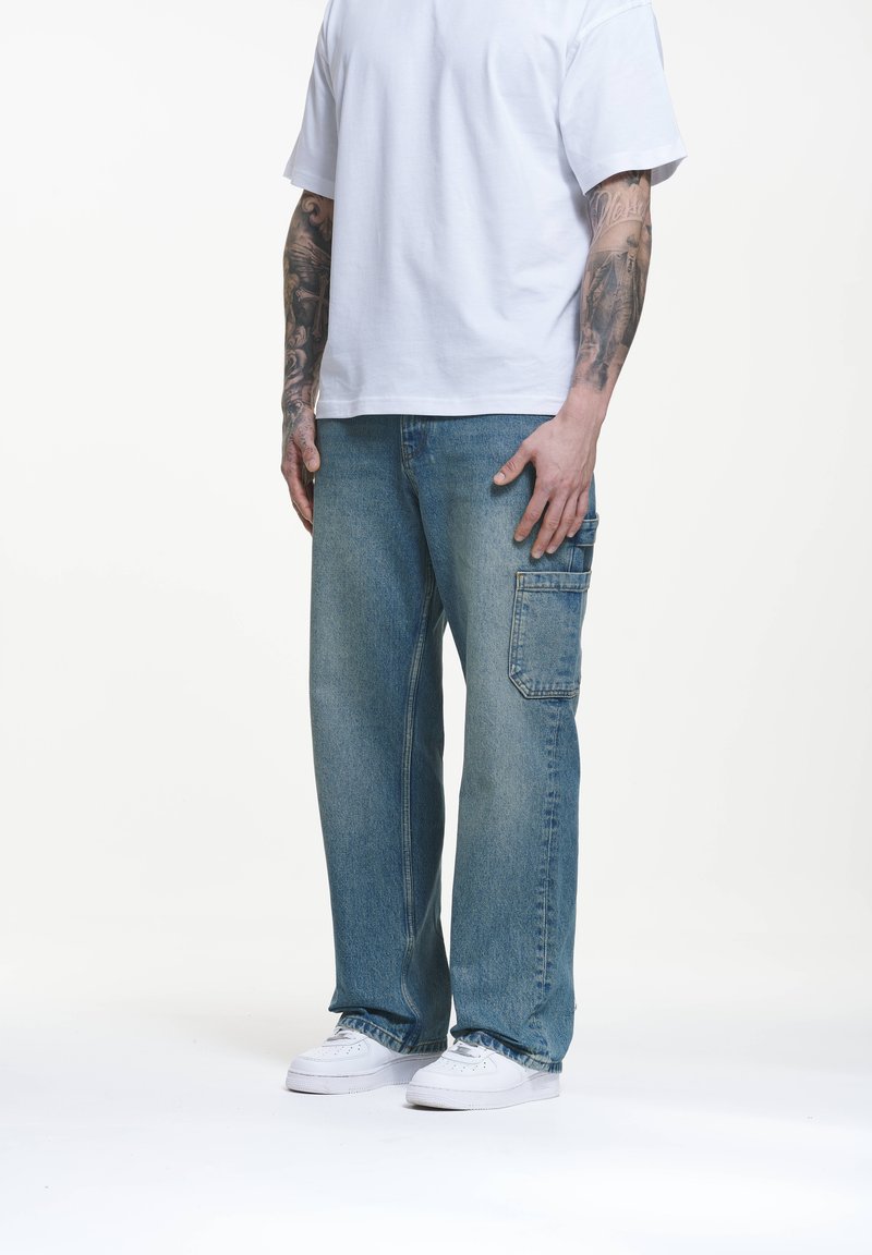 Jeans de mezclilla azul claro con un corte holgado, que cuentan con un bolsillo lateral y un ligero desgaste. Combinados con una camiseta blanca de manga corta y zapatillas blancas.