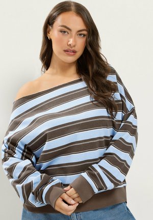 Femme aux longs cheveux marron portant un pull rayé marron et bleu épaules dénudées et un jean bleu, debout devant un fond uni.