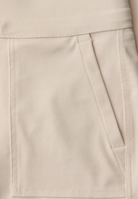 Gros plan sur un tissu beige montrant une poche avant à ouverture diagonale avec des coutures détaillées sur un pantalon ou une jupe.