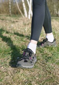 Scarpe da trekking nere e grigie con dettagli viola, suole testurizzate e lacci, indossate con calzini bianchi alla caviglia e leggings neri su prato.