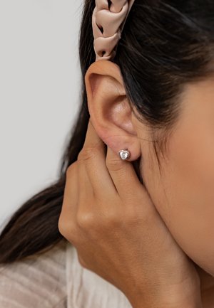 Herzförmige silberne Ohrstecker, die an einem Ohr gezeigt werden, vor einem neutralen Hintergrund mit sichtbarem Hautton und texturiertem Haar.