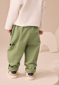 Pantalon en coton vert avec poches latérales, ourlets retroussés et coupe décontractée, associé à des baskets beiges avec un accent noir.