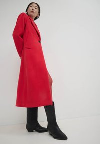 Long manteau rouge avec un col, fermeture à un bouton et deux poches latérales, associé à des bottes de cowboy noires avec un détail embossé.