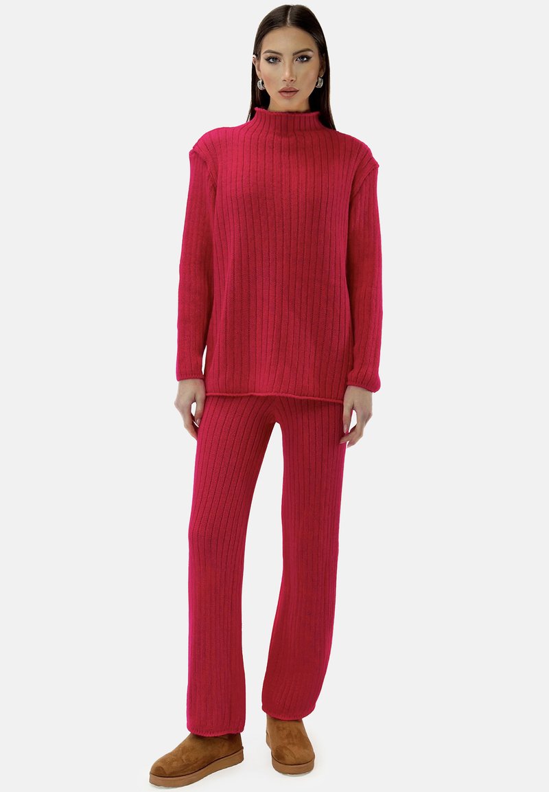 Geribbelde roze trui met een hoge hals en lange mouwen, gecombineerd met bijpassende geribbelde broek en bruine enkelboots, tentoongesteld tegen een witte achtergrond.
