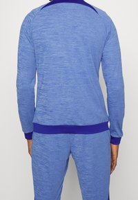 Blå atletisk træningsoverdel med tekstureret stof, kontrasterende mørkeblå krave og manchetter, lange ærmer og en tætsiddende pasform.