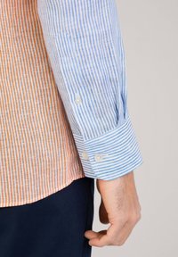 Camisa a rayas con un panel izquierdo naranja y blanco y un panel derecho azul y blanco. Cuenta con puños abotonados y un dobladillo ligeramente curvado.