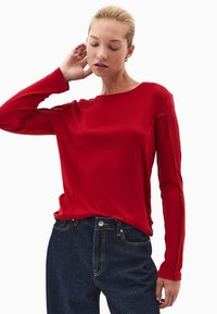 Czerwona bluzka z długim rękawem wykonana z gładkiego materiału, z okrągłym dekoltem, z detalami na szwach bocznych, zestawiona z ciemnoniebieskimi dżinsami.