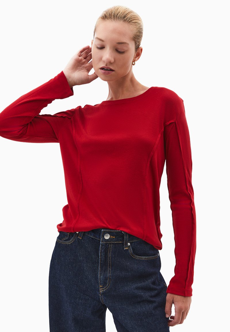 Czerwona bluzka z długim rękawem wykonana z gładkiego materiału, z okrągłym dekoltem, z detalami na szwach bocznych, zestawiona z ciemnoniebieskimi dżinsami.