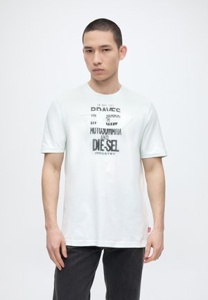 Joven con el pelo corto que lleva una camiseta blanca con texto negro y pantalones oscuros, de pie frente a un fondo claro y liso.