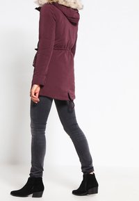 Burgundy parka med en foderhuva i päls, elastisk midja och dragkedjor, i kombination med svarta ankelstövlar och grå slim-fit jeans.