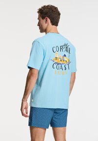 Turkizna kratka majica s kratkimi rokavi, ki prikazuje pisano plažno grafiko in besedilo "CORAL COAST SHIW". V kompletu z patterned modrimi kratkimi hlačami.