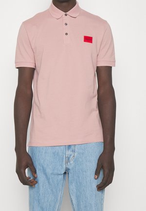 Mężczyzna ubrany w jasnoróżową koszulkę polo z czerwoną naszywką z logo Hugo na piersi oraz jasnoniebieskie dżinsy, stojący na białym tle.