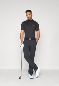 Mann i svart golfskjorte og bukser holder en kølle med en hanskebrukte hånd, stående med bena krysset mot en ensfarget bakgrunn.