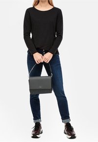 Persoon in een zwarte lange mouw shirt en blauwe jeans die met beide handen een zwarte handtas vasthoudt voor een witte achtergrond.