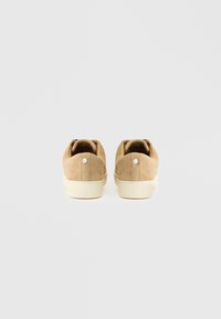 Beżowe buty zamszowe o niskim profilu, z teksturowaną powierzchnią i jasnokolorową gumową podeszwą, z małym logo z tyłu.