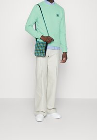 Mintgrön sweatshirt med rund halsringning, beige vida byxor och vita sneakers. Rutig axelväska i blå, grön och gula nyanser.