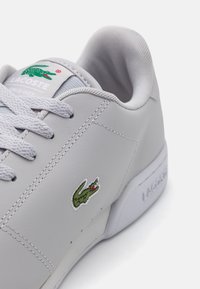 Lacoste tenisice od svijetlosivog sintetičkog kože s bijelim potplatama, sivi vezice i zelenim logom krokodila uvezenim sa strane.