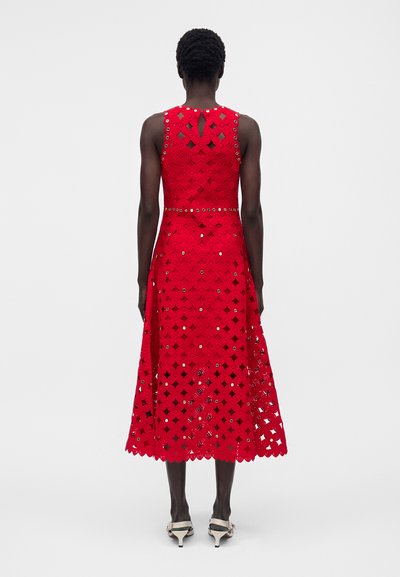 Γυναίκα με κοντά σγουρά μαλλιά που φοράει μια κόκκινη δαντελωτή midi φόρεμα χωρίς μανίκια με κυκλικές ανοίγματα και ασημένιες λεπτομέρειες, συνδυασμένο με λευκά πέδιλα με τακούνι.