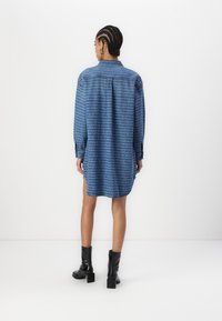 Blaue Denim-Shirtkleid mit langen Ärmeln, Rückenschlitzen, seitlichen Schlitzen und einem sich wiederholenden Textmuster. Kombiniert mit schwarzen Knöchelstiefeln.