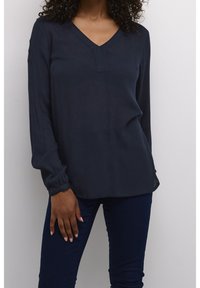 Blouse bleu marine à manches longues avec un col en V et un tissu subtilement texturé. Présente une coupe ample et des poignets froncés. Portée avec un jean foncé.