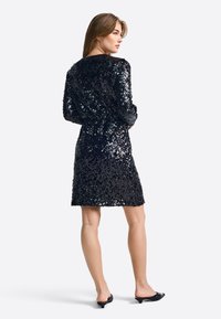 Crna haljina sa šljokicama, dugih rukava, do koljena i s okruglim izrezom. Uparena s crnim potpeticama. Glatka, sjajna tekstura.