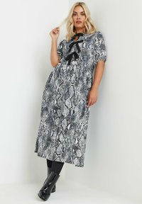 Mekko, jossa on musta-valkoinen käärmeennahkakuosi, rusetti kaula-aukossa, lyhyet puffihihat ja rento siluetti, yhdistettynä kiiltäviin mustiin nilkkasaappaisiin.