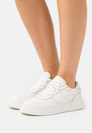 Jambes portant des baskets blanches basses à lacets avec des semelles épaisses, debout sur une surface blanche unie.