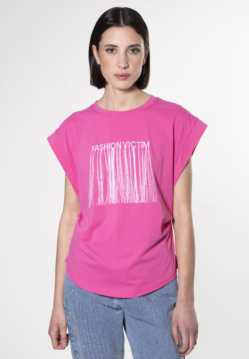 Lyserød t-shirt med korte ærmer og "FASHION VICTIM" trykt i hvid tekst samt frynsede detaljer nederst. Parret med glitrende blå jeans.