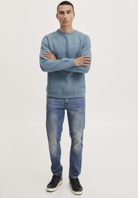 Lichtblauwe sweatshirt met ronde hals en ribgebreide mouwen, gecombineerd met vervaagde blauwe jeans en zwarte sneakers, staande tegen een effen achtergrond.
