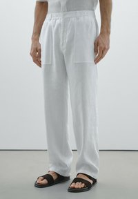 Pantalons en lin blancs avec une coupe décontractée, dotés de deux grandes poches avant et d'une fermeture à bouton à la taille. Sandales noires avec une lanière.