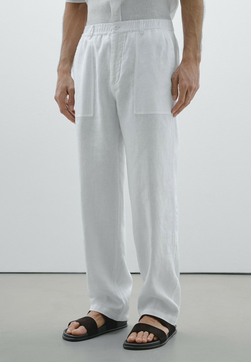 Pantalons en lin blancs avec une coupe décontractée, dotés de deux grandes poches avant et d'une fermeture à bouton à la taille. Sandales noires avec une lanière.