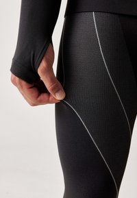 Zwarte sportlegging met een gestructureerde afwerking, voorzien van witte stiksels in een diagonale patroon en een strakke pasvorm. Een hand houdt de stof vast.
