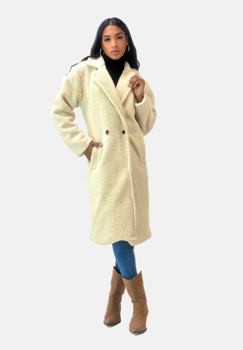 Abrigo de felpa beige de doble botonadura con cuello de solapa, bolsillos de parche y textura suave, llevado sobre un suéter negro de cuello alto y jeans azules.